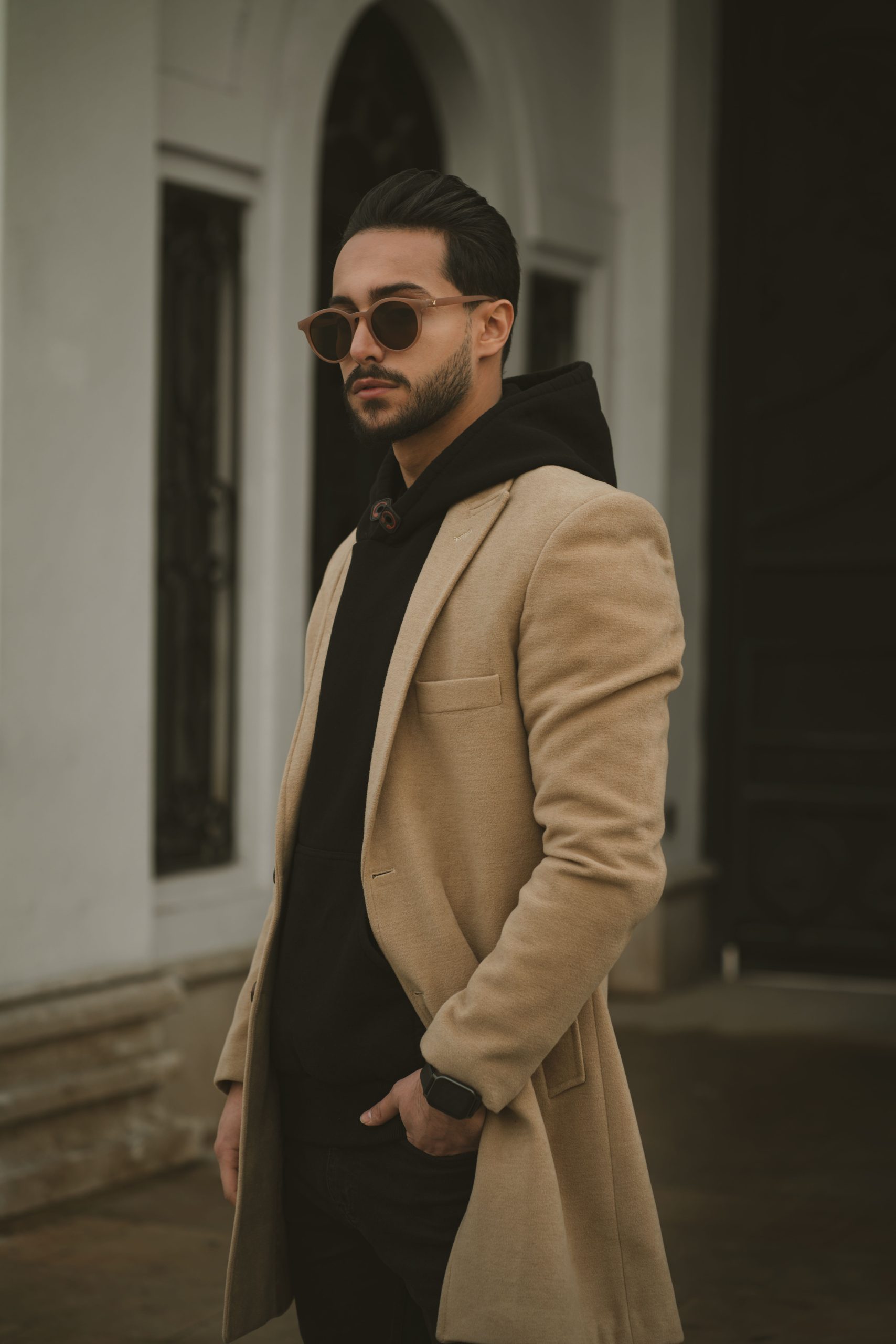 Man in beige coat posing outside an urban building, showcasing trendy fashion style.