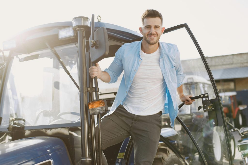 Man smiling and standing by a tractor in sunlight, wearing casual blue attire.