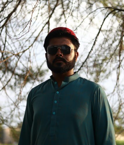 A striking portrait of a man wearing traditional clothing and sunglasses, standing outdoors in nature.