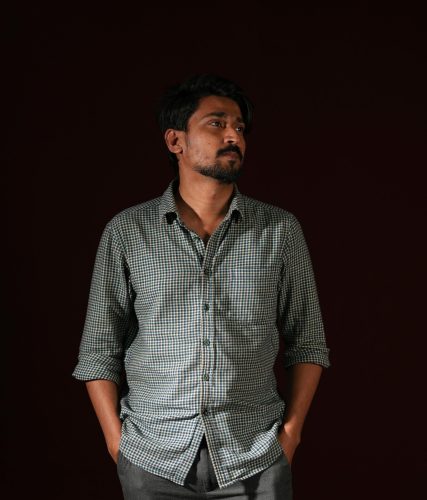 Contemplative man in checkered shirt stands against a dark background, hands in pockets.